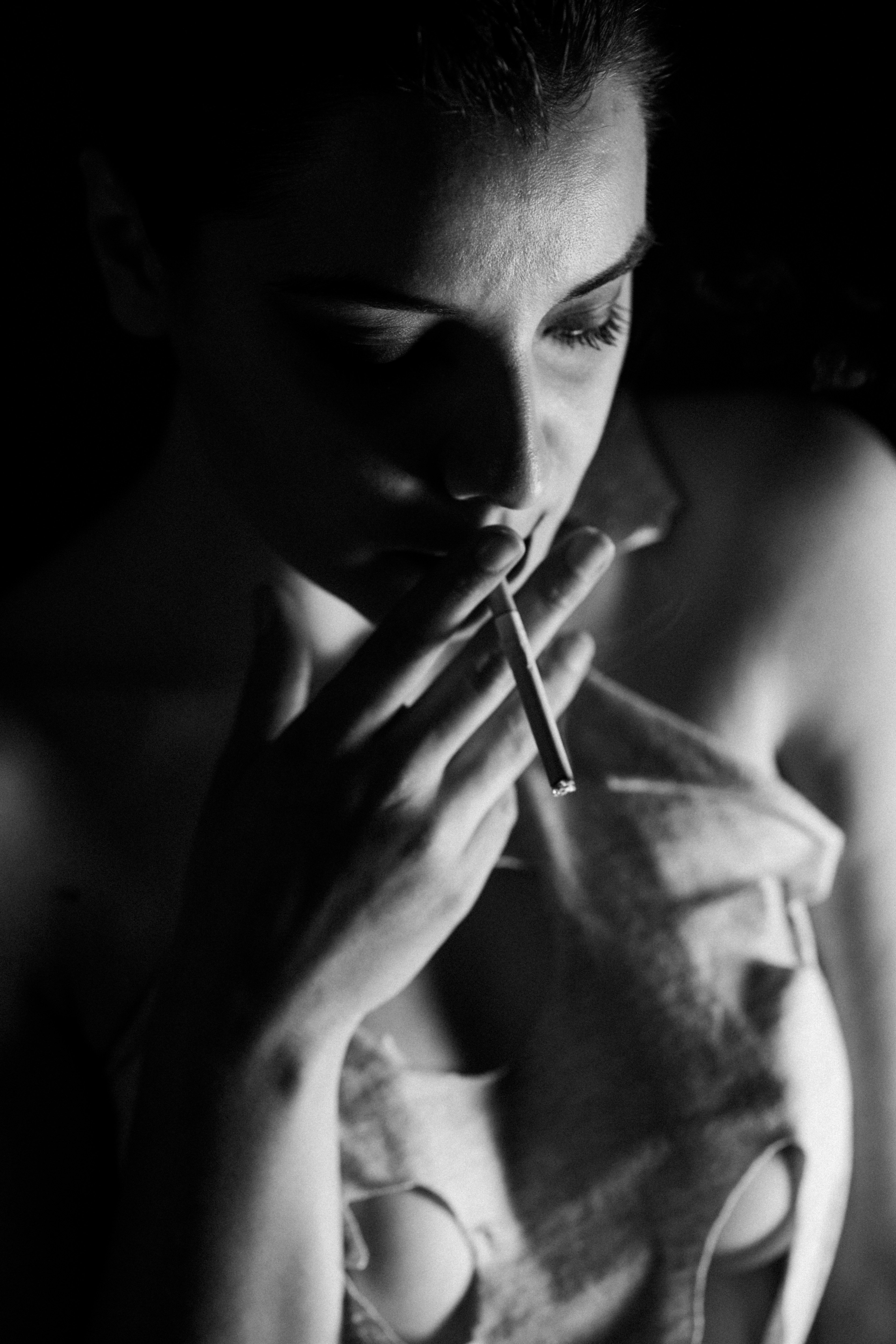 Primo piano in bianco e nero del busto di una donna a petto nudo; mano destra con sigaretta vicino alle labbra, testa leggermente china, luce laterale forte, sfondo scuro.