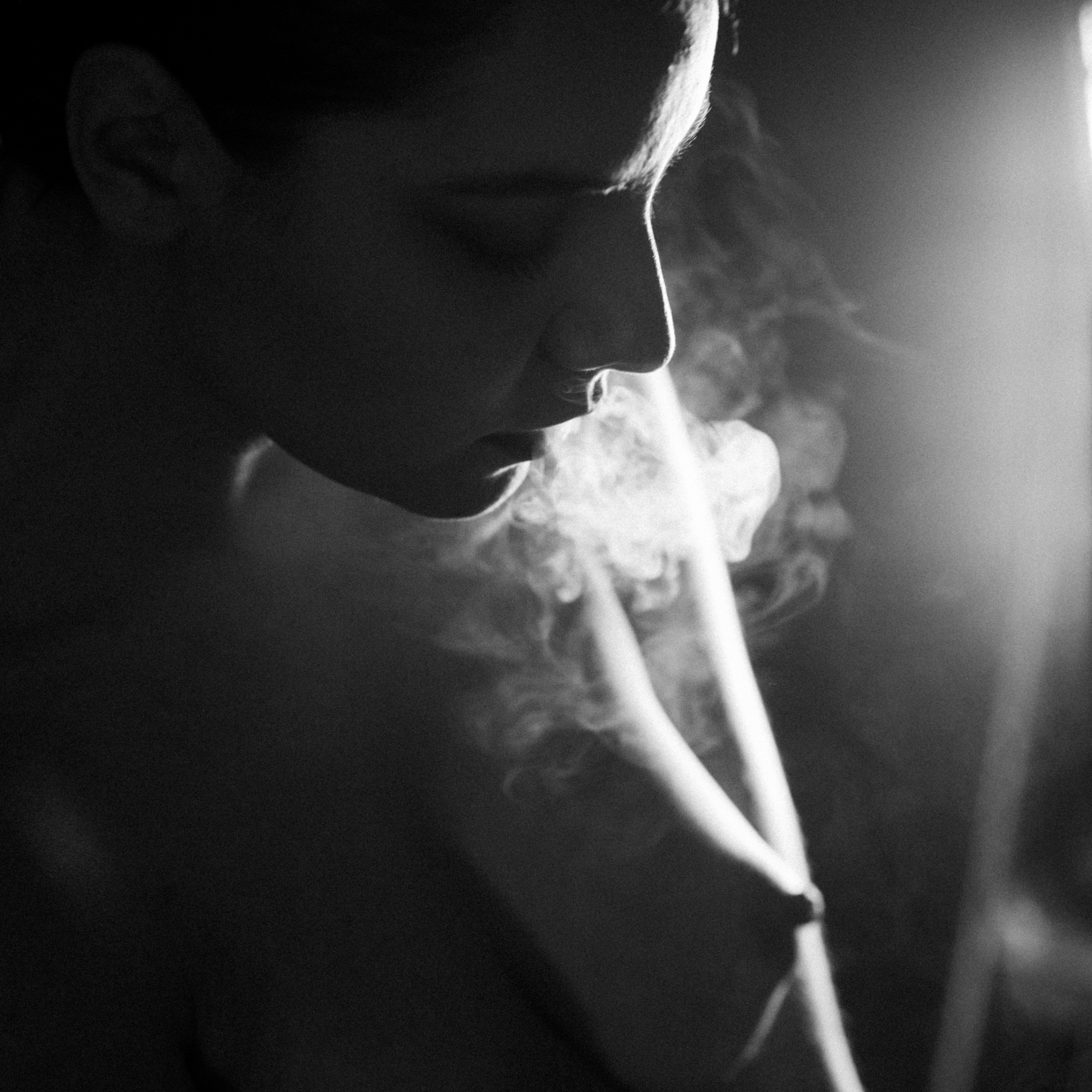 Profilo in bianco e nero di una donna che espira fumo; spalla e collo scoperti, braccio piegato, fascio di luce laterale su viso e fumo, fondo scuro.