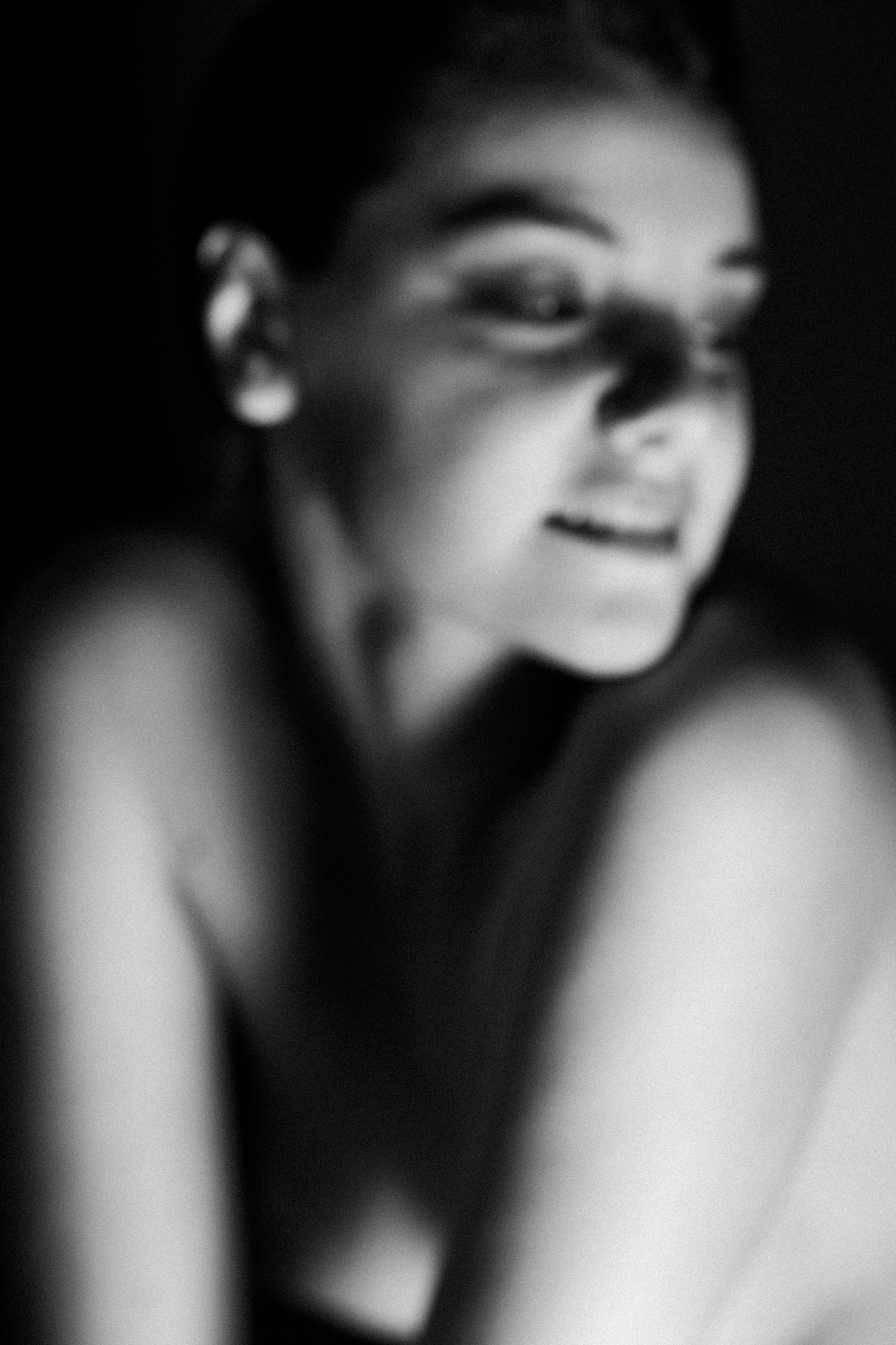 Primo piano sfocato in bianco e nero del volto; donna inclinata in avanti, occhi mezzo chiusi, spalle nude, luce morbida che modella guancia e naso.