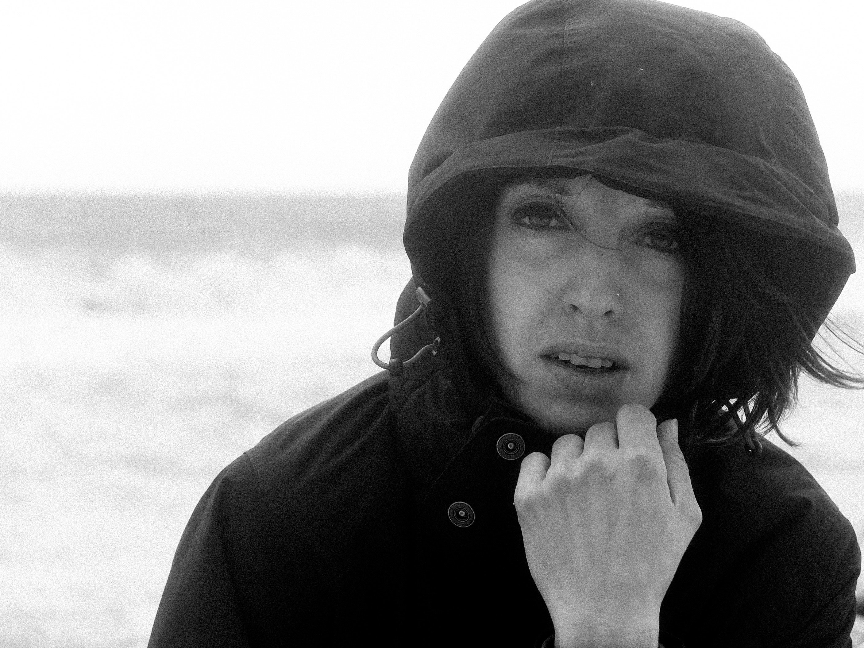 Ritratto all’aperto vicino al mare; donna con giacca scura e cappuccio, mano alla bocca, vento nei capelli, orizzonte e onde sullo sfondo.