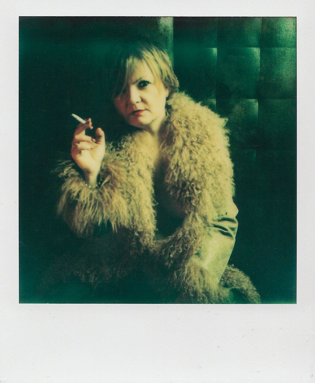 Immagine in stile Polaroid: donna seduta su divano verde, cappotto peloso chiaro, gambe accavallate, sigaretta in mano, luce diretta da flash.