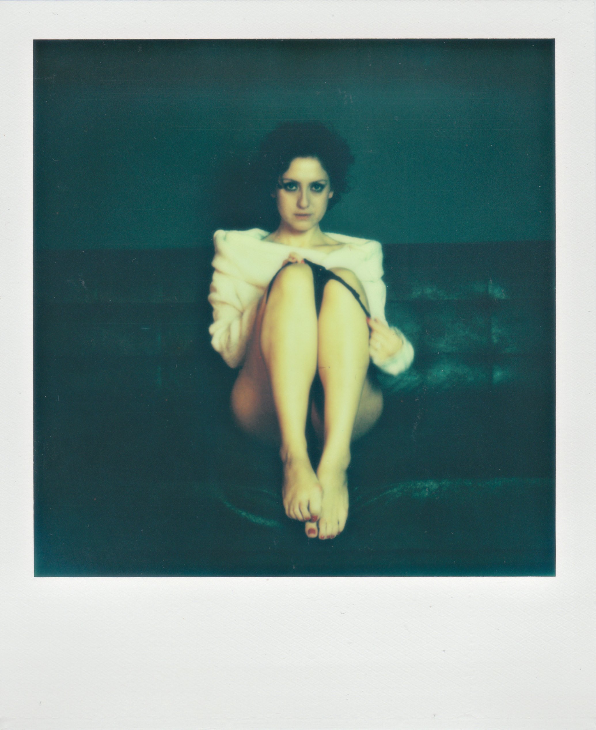 Scatto in stile Polaroid su sfondo verde; donna seduta con spalle scoperte, ginocchia raccolte al petto e braccia intorno alle gambe, piedi nudi.