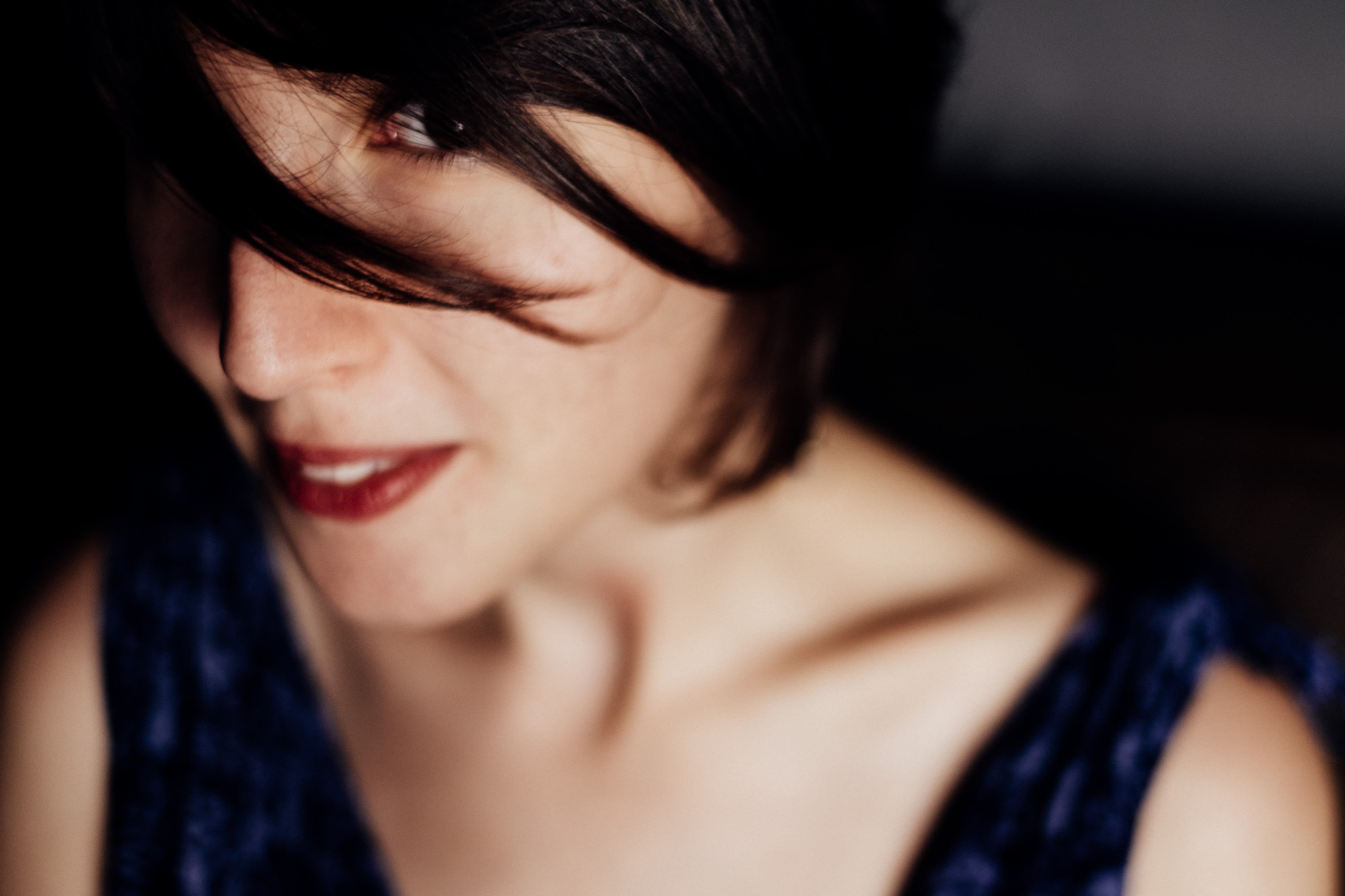 Primo piano obliquo del volto con sorriso accennato; spalle scoperte, spallina blu visibile, capelli ricadono su un lato, luce calda.