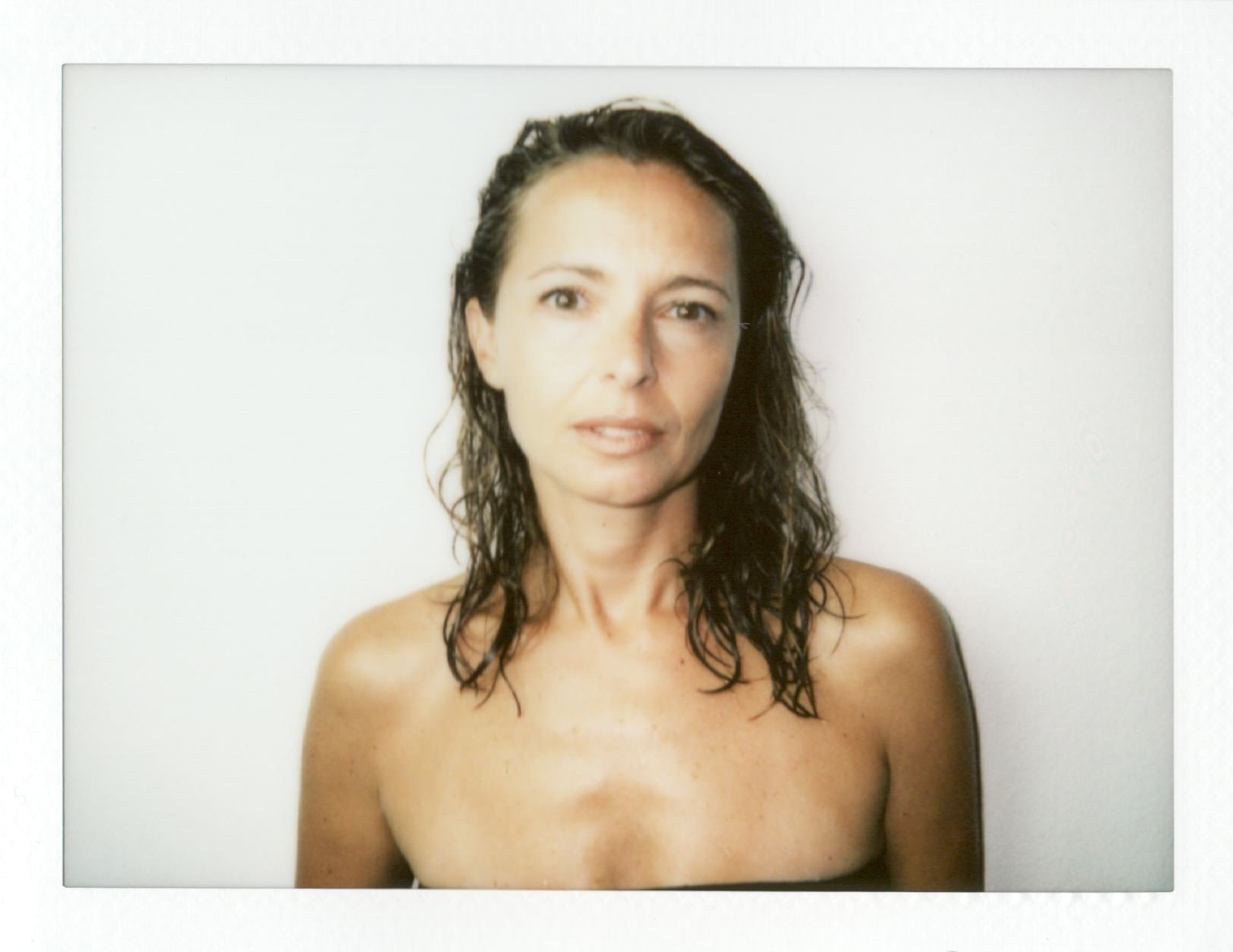 Immagine in stile Polaroid: donna a seno nudo fino all'altezza del busto, capelli bagnati o spettinati che cadono sulle spalle, sguardo diretto verso l'obiettivo con espressione neutra; bordo bianco tipico della Polaroid visibile attorno alla fotografia; luce da flash diretta che crea ombre nette dietro il soggetto.
