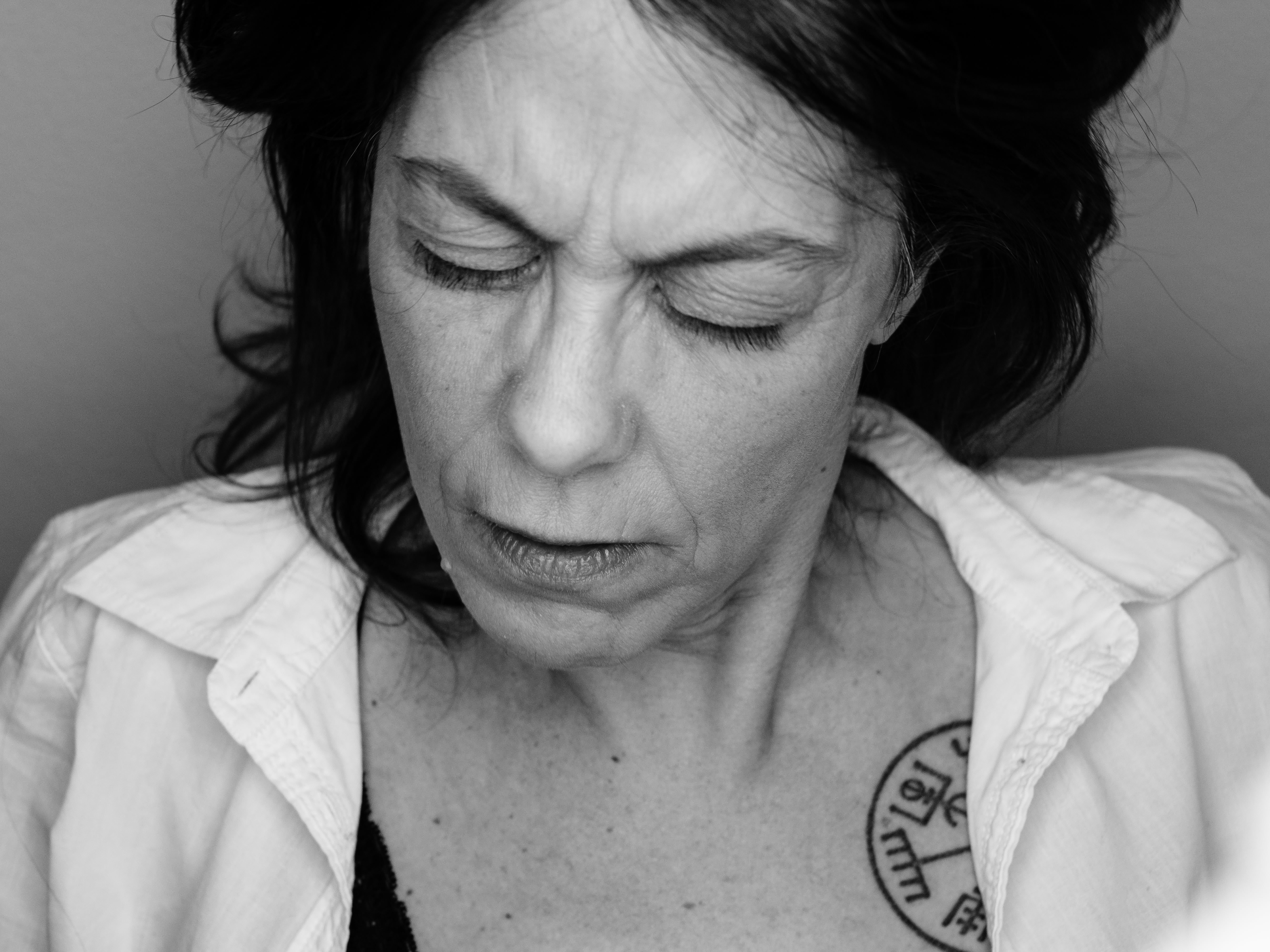 Primo piano in bianco e nero di una donna con espressione concentrata; capelli scuri, sguardo inclinato verso il basso, camicia aperta che mostra parte della spalla e un tatuaggio circolare sul petto; luce laterale che evidenzia la texture della pelle.