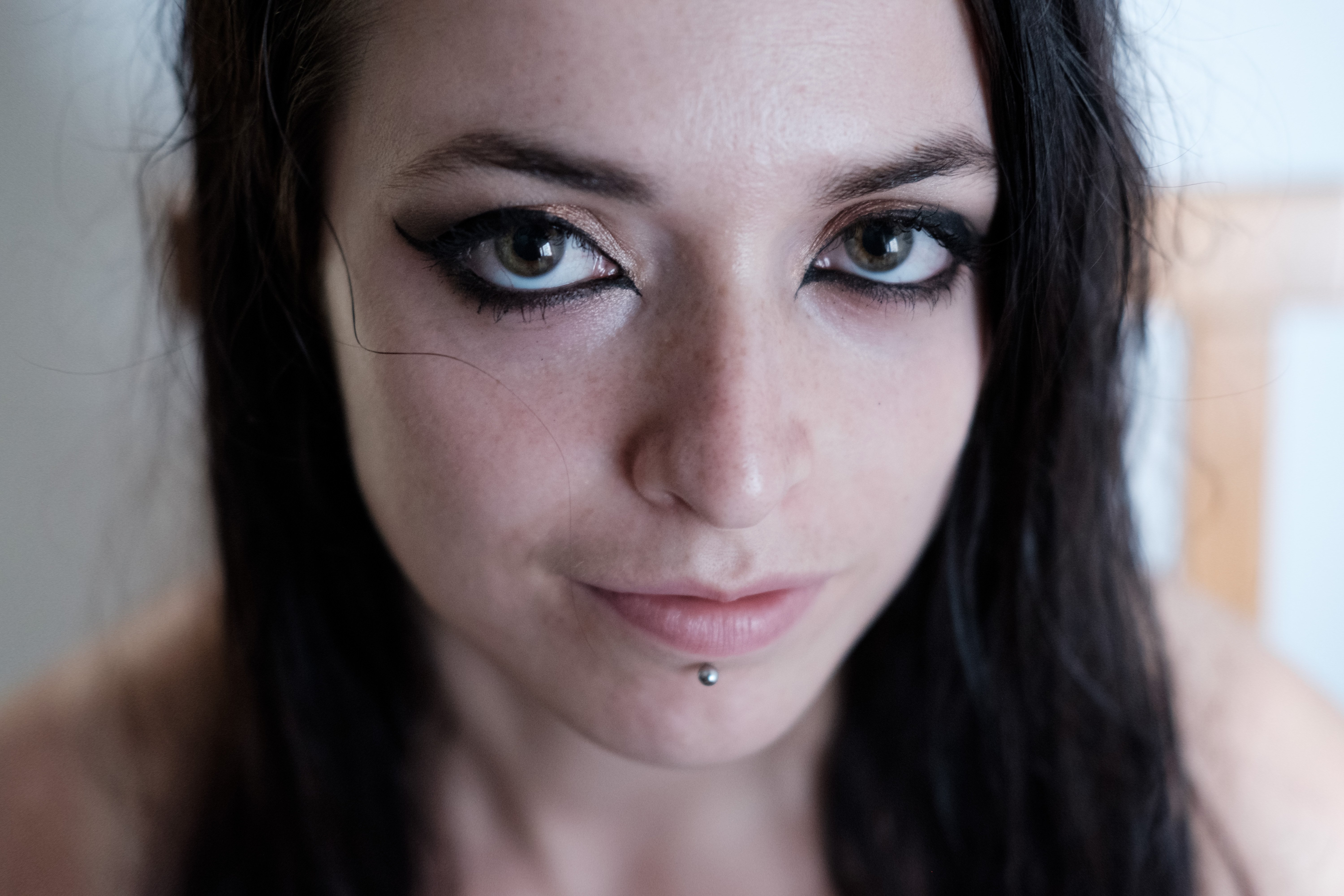 Color close-up of the face with light makeup; eyes emphasized by dark eyeliner, lower lip pierced with a stud, neutral expression, straight dark hair over the shoulders; blurred indoor background.