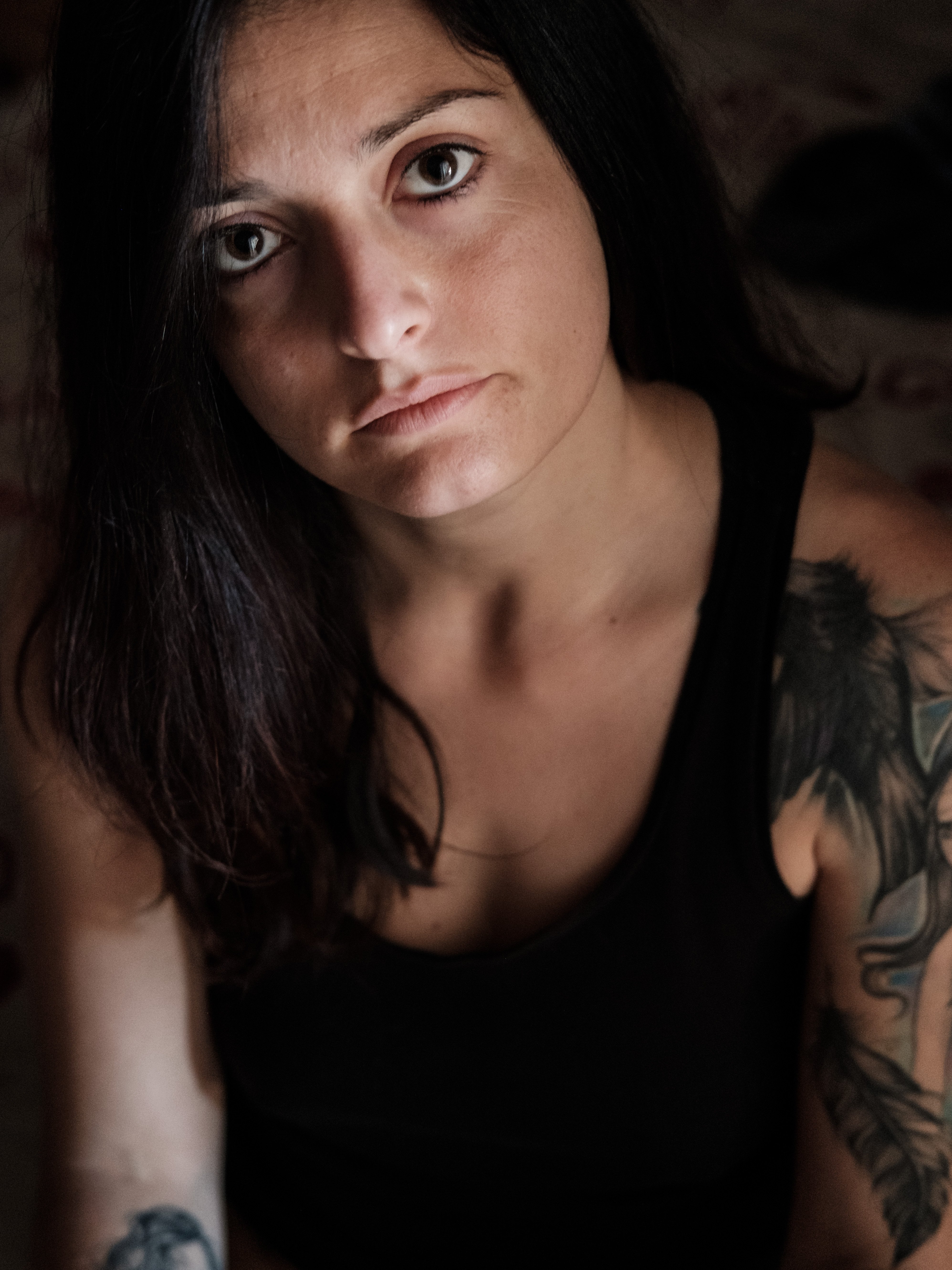 Color close-up of a young woman with dark eyes and pronounced makeup; direct gaze at the camera, lower lip pierced, long dark hair; soft natural light, blurred indoor background.
