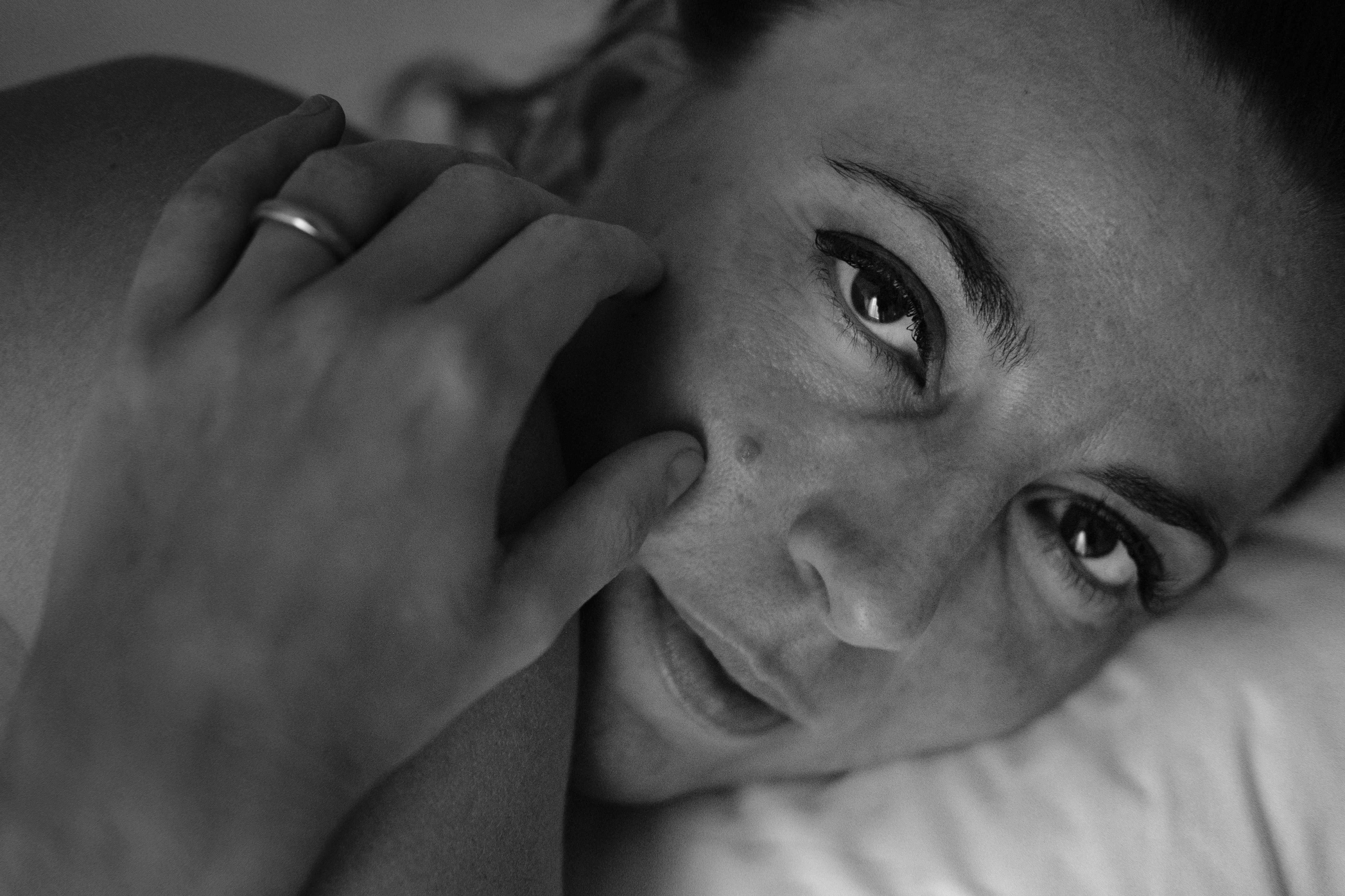 Primo piano in bianco e nero di una donna sdraiata su un cuscino: il volto riempie l’inquadratura, occhi aperti che guardano in camera; mano vicino al viso con le dita che sfiorano la guancia; capelli scuri tirati all’indietro; luce laterale morbida che modella sopracciglia, naso e labbra.