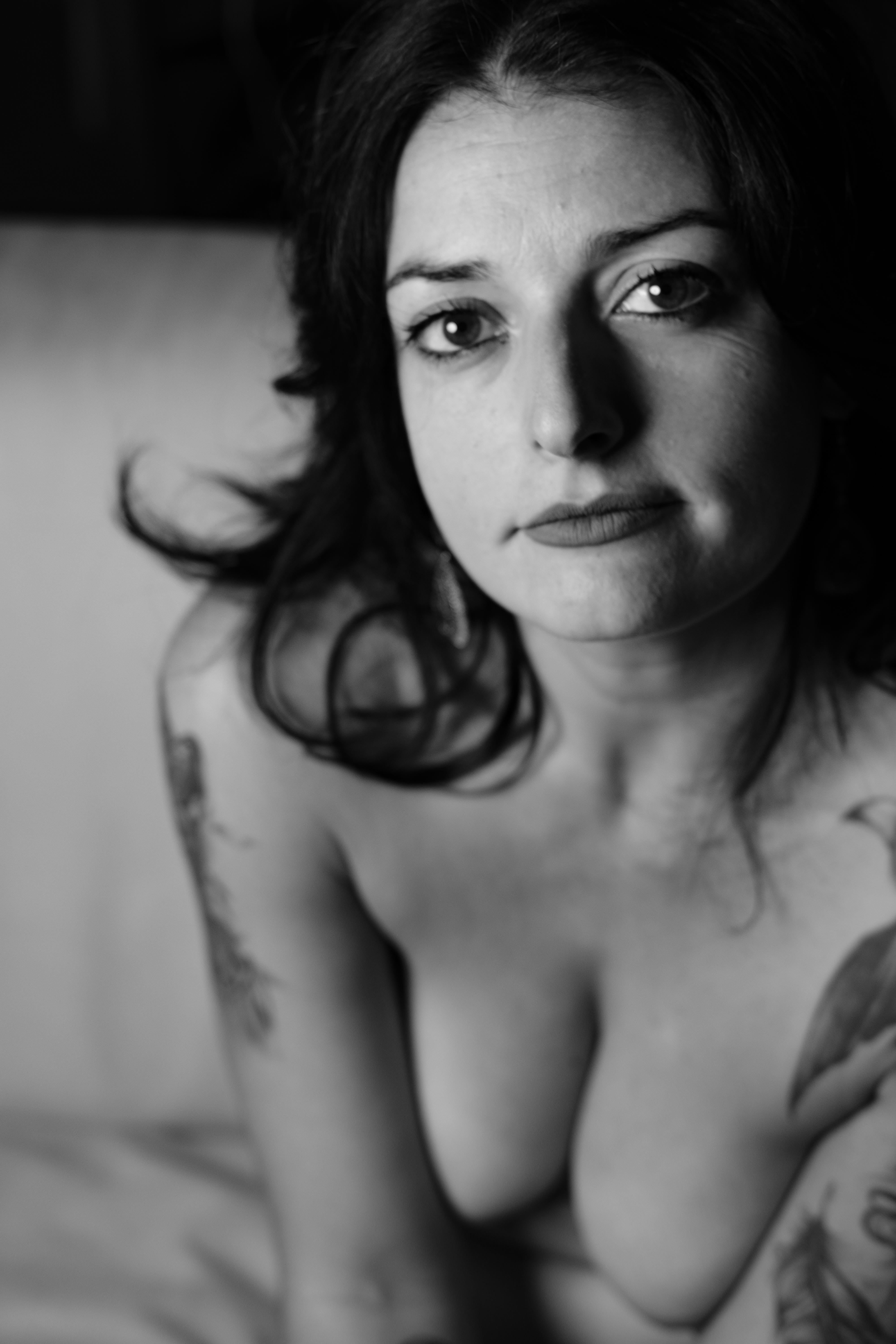Black‑and‑white tight portrait: woman seated on a couch, bare shoulders and breasts in the extreme foreground; frontal face with direct gaze and light lipstick; short dark hair; soft light drawing collarbones, breasts and facial features.